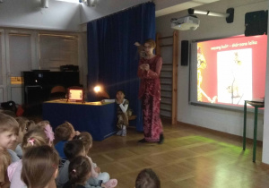 dzieci siedzą na ławeczkach, Mama dziewczynki, ubrana w kobiecy strój indonezyjski opowiada dzieciom o wyspie Jawa i teatrze cieni, w tle prezentacja multimedialna