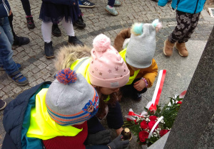 dwie dziewczynki i chłpoiec stawiają zapalony znicz pod pomnikiem Marszałka J. Piłsudskiego