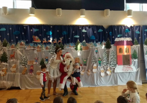 Mikołaj trzyma dziewczynkę na kolanach, Śniezynka stoi obok i trzyma chłopca za rękę