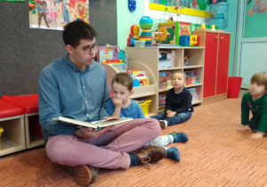 Tata czyta dzieciom książkę, syn siedzi obok , reszta dzieci siedzi i słucha