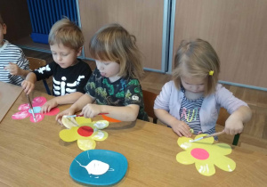 Dzieci siedzą przy stole i smarują sylwety kwiatów klejem