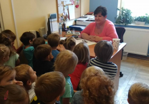 Dzieci stoją w gabinecie i słuchają pani Kierownik która siedzi za biurkiem i opowiada o swojej pracy