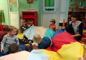 Kilkoro dzieci siedzi na kolorowej chuście , reszta dzieci rytmicznie porusza chustą