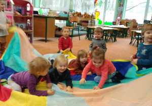 Kilkoro dzieci siedzi na kolorowej chuście , reszta dzieci rytmicznie porusza chustą