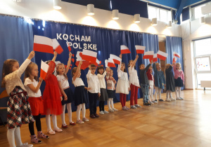 Dzieci ubrane na białoczerwono stoją na scenie i machają białoczerwonymi flagami