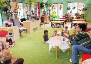 Nauczycielka przeprowadza eksperyment a dzieci siedza na dywanie i obserwują zachodzace zmiany