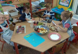 Dzieci siedzą przy stolikach i kroją owoce do sałatki owocowej
