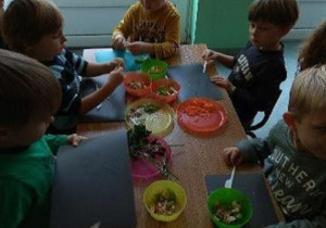 Dzieci siedzą przy stolikach i kroją składniki sałatki z ugotowanej fasoli, jabłka, ziaren słonecznika, jabłka